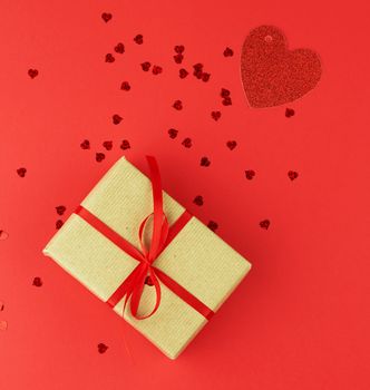 rectangular box wrapped in brown kraft paper and tied with red thin silk ribbon in red, decorative shiny hearts are scattered nearby, holiday is Valentine's Day. Anniversary, birthday. 