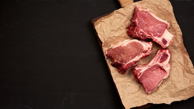 three raw juicy pork slices of meat on the rib, food lies on brown parchment paper,  black wooden background and an empty space on the left for writing a recipe or menu, top view