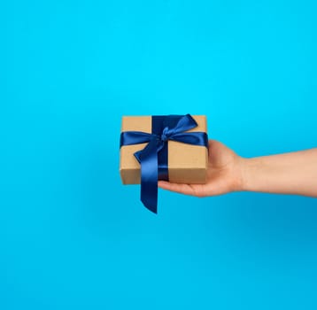 hand holds a box with a gift wrapped in brown kraft paper and tied with a silk blue ribbon on a blue background, concept of giving a gift