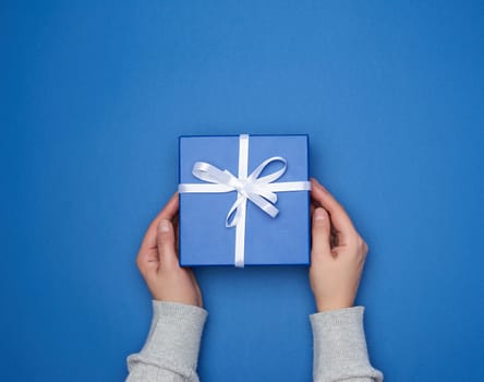 two female hands in a sweater hold a square blue box with a bow on a blue classic background, top view, festive backdrop