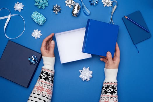 two female hands in a sweater hold a square blue box with a bow on a blue classic background, top view, festive backdrop