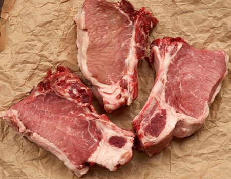 three raw juicy pork slices of meat on the rib, food lies on brown parchment paper,  black wooden background, top view