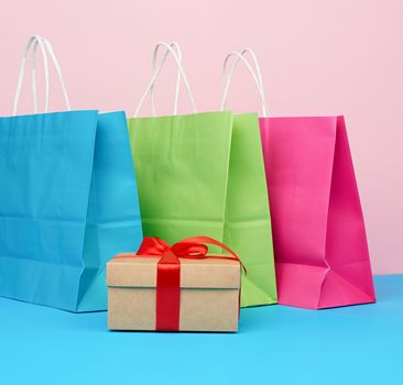 brown paper box with a gift and multi-colored paper bags for shopping and gifts with white handles stand on a pink-blue background
