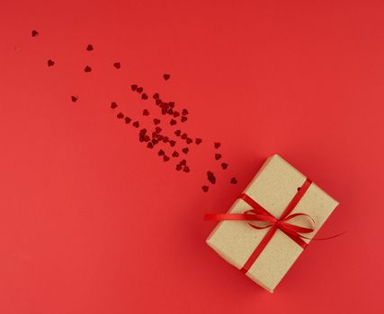box wrapped in brown kraft paper and tied with a red thin silk ribbon, scattered confetti in the shape of hearts, red background, festive backdrop for Valentine's Day