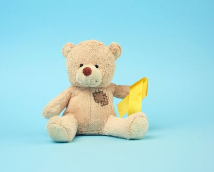 Cute brown teddy bear with a patch holds a silk yellow ribbon in the shape of a loop on a blue background, concept of the fight against childhood cancer, problem of suicides and their prevention