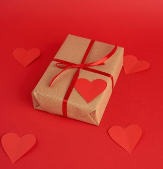 box wrapped in brown kraft paper and tied with a red thin silk ribbon, cut out of paper hearts, red background, gift for Valentine's Day, birthday