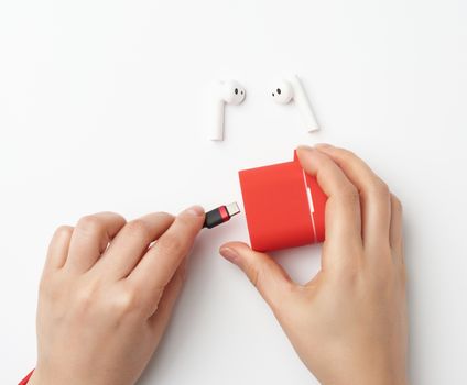 two female hands are holding a cable and a red box with wireless headphones, white table, top view