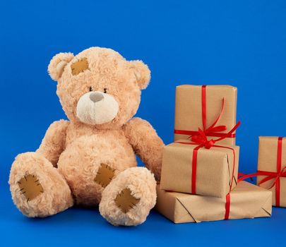 big beige teddy bear with patches, stack of gifts in boxes wrapped in brown paper, tied with silk ribbon on a blue background, happy birthday and valentines day
