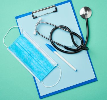 metal stethoscope, paper holder with a blank blue sheet, disposable blue medical mask on a green background, top view, flat lay