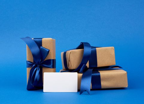 empty white paper business card and a stack of boxes wrapped in brown kraft paper and tied with blue silk ribbon, gifts lie on a dark blue background