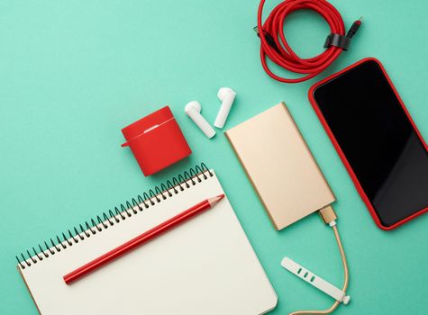 paper notebook, power bank with cable, red smartphone with blank black empty screen, headphones on green background, workplace, top view
