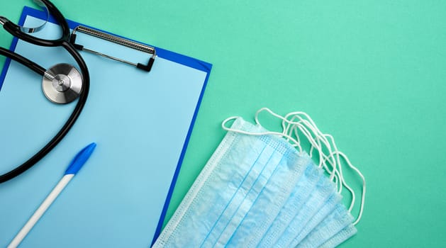 metal stethoscope, paper holder with a blank blue sheet, disposable blue medical mask on a green background, top view, flat lay