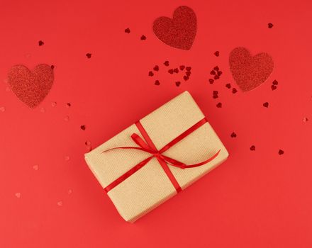 box wrapped in brown kraft paper and tied with a red thin silk ribbon, scattered confetti in the shape of hearts, red background, festive backdrop for Valentine's Day