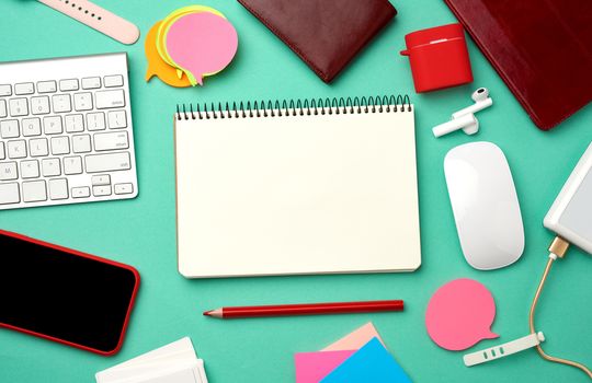 paper notebook, keyboard, power bank with cable, red smartphone with blank black empty screen, headphones, blank paper business cards and wireless mouse on green background, workplace, top view