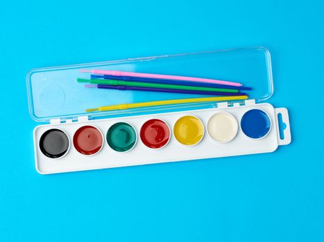 multicolored watercolor paints in a plastic box and brushes on a blue background, set for drawing, close up