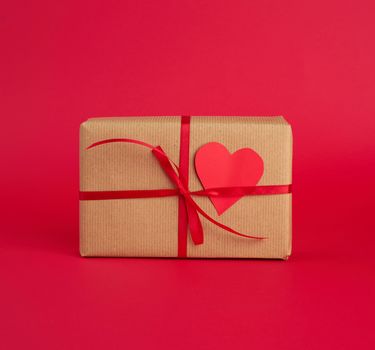 box wrapped in brown kraft paper and tied with a red thin silk ribbon, cut out of paper hearts, red background, gift for Valentine's Day, birthday, flat lay