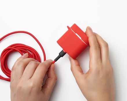 two female hands are holding a cable and a red box with wireless headphones, white table, top view