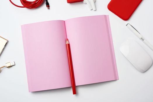 open pink paper notebook, power bank with cable, red smartphone, headphones, wireless mouse on white background, workplace, top view, flat lay