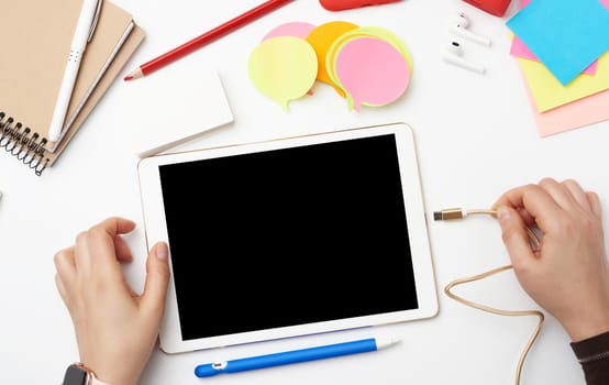white electronic tablet and two hands, the process of working or drawing on the screen, a workplace with a notebook, headphones and color stickers, top view