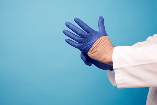 male doctor in white uniform puts on his hands blue sterile latex gloves before surgery, blue background, copy space