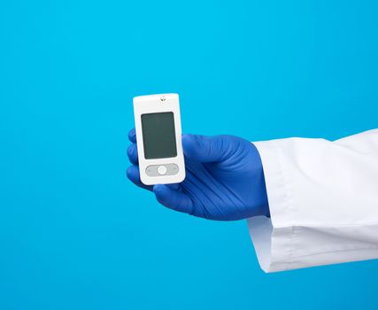 male hand with blue sterile gloves holds a white blood glucose meter for measuring blood sugar, blue background