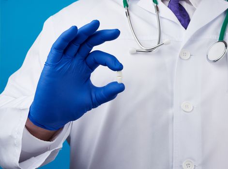 male hand in blue sterile gloves holds an oval white pill, medical treatment concept for diseases, blue background