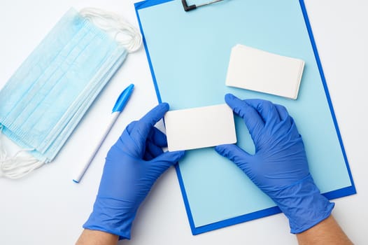 two hands in blue medical gloves are holding rectangular white blank business cards, top view, white background