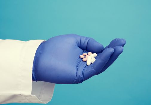 male hand with blue sterile gloves holds white pills, concept of drug treatment for diseases, blue background