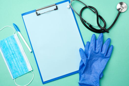 metal stethoscope, paper holder with a blank blue sheet, disposable blue medical mask on a green background, top view, flat lay