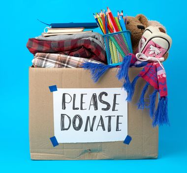 brown cardboard box with a white sheet pasted and the inscription please donate on a blue background, inside clothes, toys, stationery, help concept
