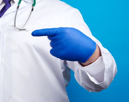 medic man in white coat with buttons, on hands wearing blue sterile gloves, showing hand gesture indicating the subject, blue background