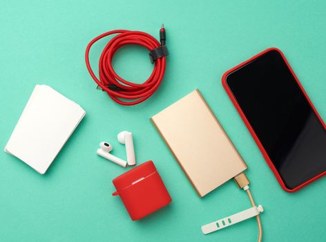 golden power bank with cable, red smartphone with blank black screen, box with headphones, empty paper business cards on green background, top view