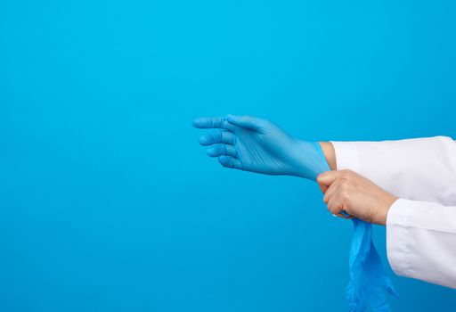female doctor in a white coat puts on her hands blue latex sterile medical gloves on her hands, blue background, place for text
