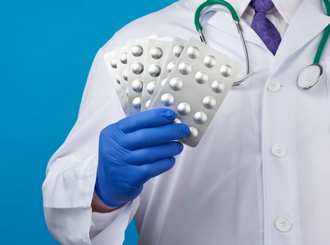 male hand with blue sterile gloves holds a stack of pills in a blister, concept for drug treatment of diseases, blue background
