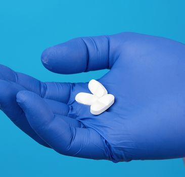 male hand with blue sterile gloves holds white pills, concept of drug treatment for diseases, blue background, close up