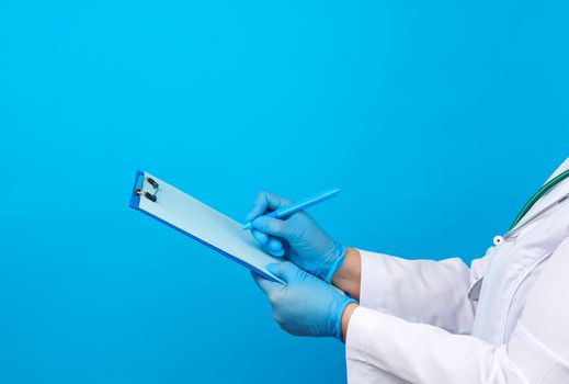 female doctor in a white coat, rubber blue medical gloves holds a folder for papers and with his right hand writes on a sheet, blue background