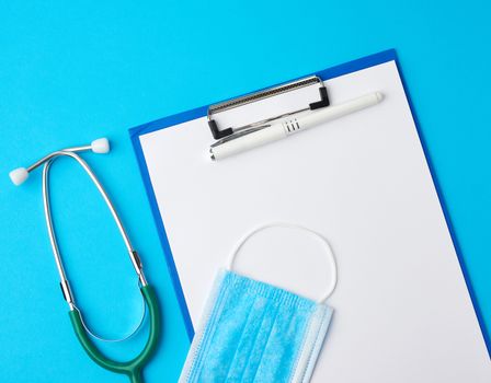 blank white sheet of paper and blue viscose medical mask for protection against the virus, green stethoscope, blue background