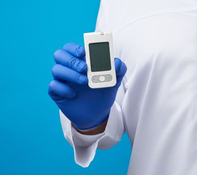 male hand with blue sterile gloves holds a white blood glucose meter for measuring blood sugar, blue background