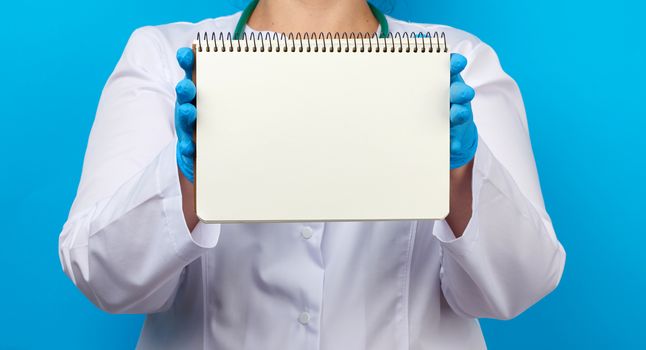medic woman in white coat, blue latex gloves holding an open notebook with blank white sheets, blue background, place for text