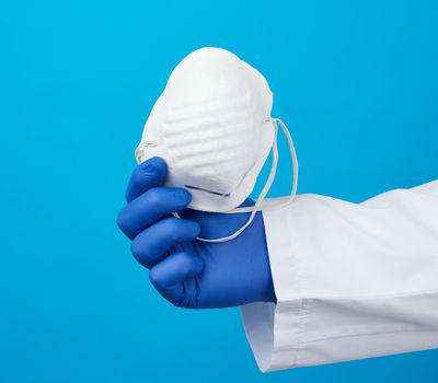 doctor in a white coat, blue latex sterile gloves holds textile disposable  masks in his hand on a blue background, additional protection against viruses during an epidemic, pandemic