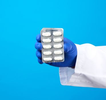 hand with blue sterile gloves holds a pills in a blister, concept for drug treatment of diseases, blue background