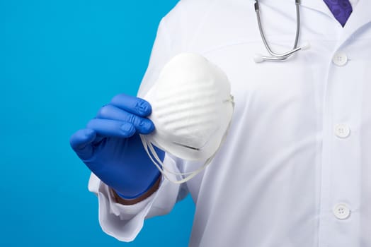 male doctor in blue latex sterile gloves holds a white disposable mask made of non-woven material on a blue background, protective accessory for the respiratory tract from the virus
