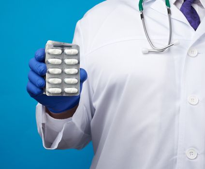 male hand with blue sterile gloves holds a stack of pills in a blister, concept for drug treatment of diseases, blue background