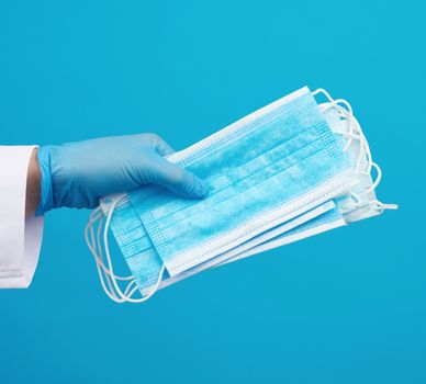 doctor in a white coat, blue latex sterile gloves holds textile disposable medical masks in his hand on a blue background, protective accessory for the respiratory tract from the virus