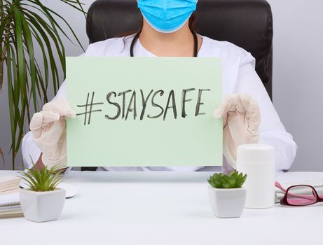woman doctor in a white coat, sterile medical gloves holds a poster with the inscription stay safe, concept of self-isolation during a pandemic and epidemic