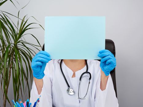 doctor in a white medical coat is sitting at a table in a brown leather chair and holding a empty blue sheet of paper in his hands, place for an inscription, medical office