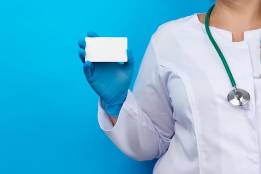 female doctor hand in blue medical latex gloves holds an empty paper business card on a blue background, place for an inscription