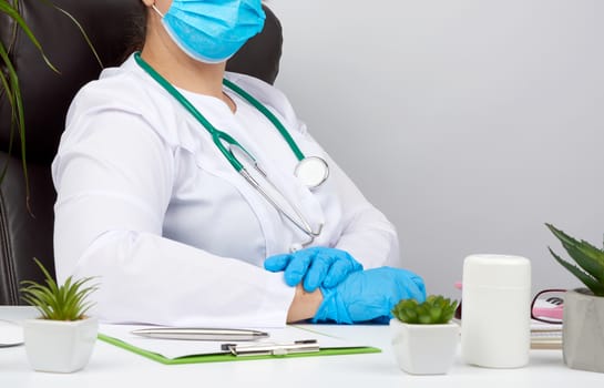 doctor in a white coat and blue latex gloves sits at a white work table in his office, concept of patient reception in the clinic