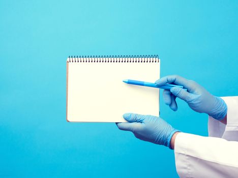 medic woman in white coat, blue latex gloves holding an open notebook with blank white sheets, blue background, place for text