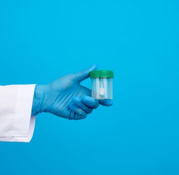 doctor in a white coat and blue sterile latex gloves holds a plastic jar for stool analysis, blue background